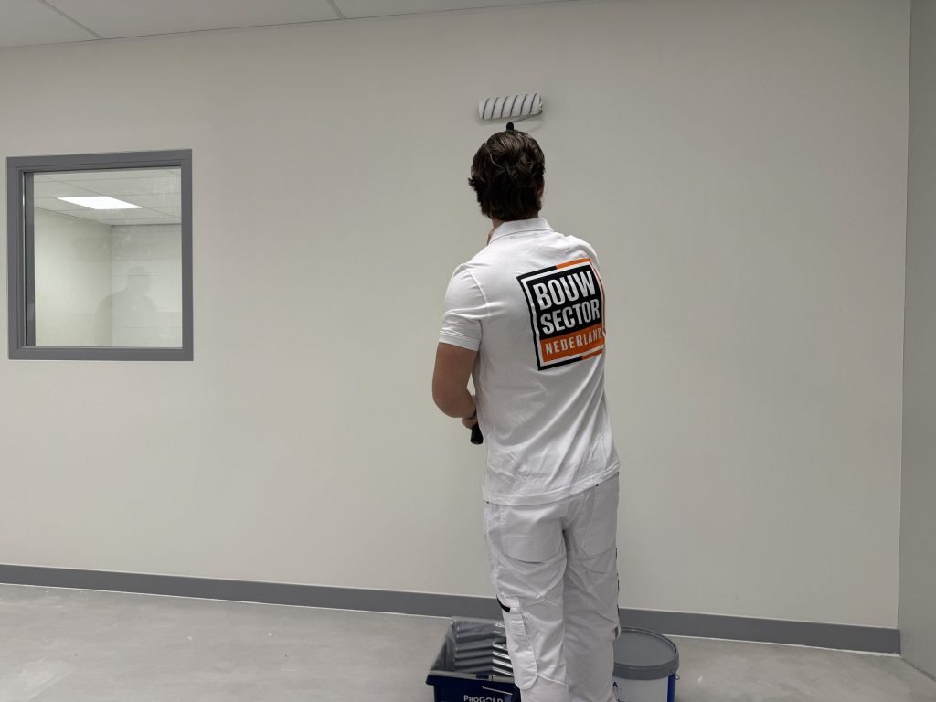 Schilder in wit werkpak rolt een witte wand, met verfbak en emmer aan zijn voeten