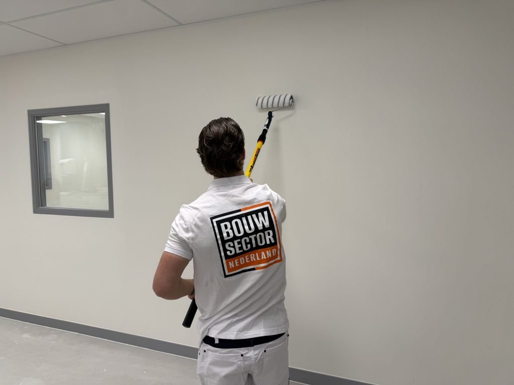 Schilder rolt verf op een witte muur naast een binnenraam met een grijs kozijn