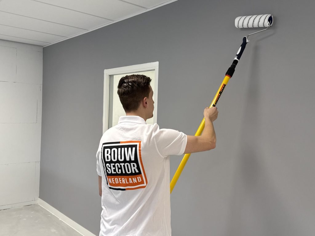 Schilder van Bouwsector Nederland rolt een grijze muur met een roller aan een verlengsteel, naast een binnenraam