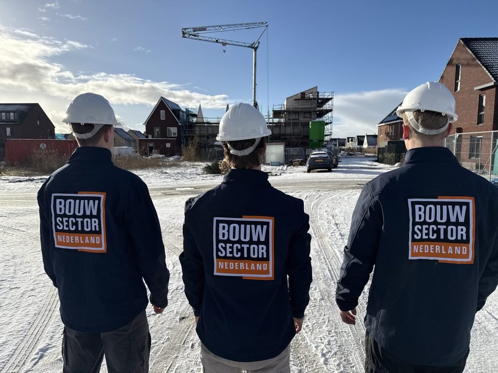Drie vakmensen van Bouwsector Nederland in donkere jas met logo en witte helm, op een besneeuwde weg tussen bakstenen woningen