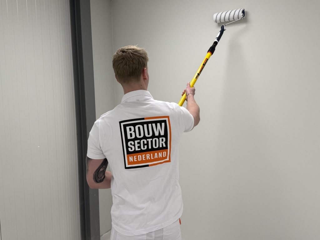 Schilder met het Bouwsector-logo op zijn rug rolt met een verlengsteel een lichte wand naast een deurkozijn